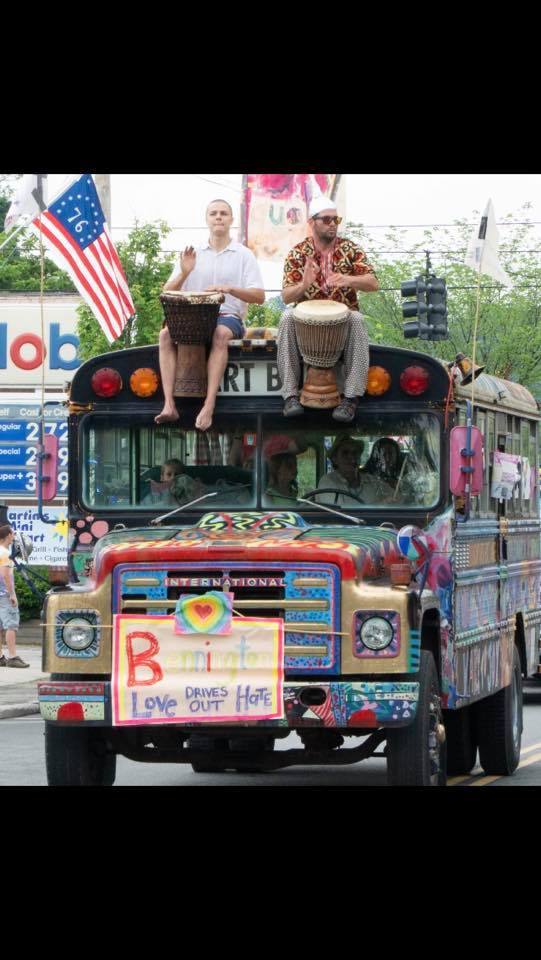 The Vermont Arts Exchange art bus drives in a pride parade. Two people sit atop the bus playing drums. The bus has a poster on its hood which reads "Bennington Love Drives Out Hate."