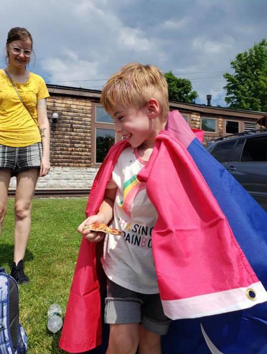 Photo of a young child wearing a bisexual pride flag as a cape. The child is smiling and holding a piece of pizza, while a smiling adult looks on from the background.
